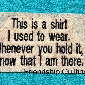 May include: A beige fabric square with black embroidered text that reads: "This is a shirt I used to wear. Whenever you hold it, know that I am there." The bottom of the square has the text "Friendship Quilting" in black.