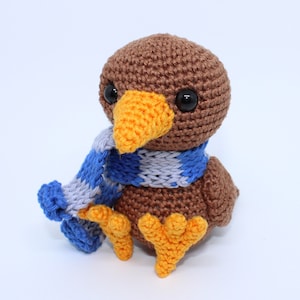 Pode incluir: Um pássaro de crochê marrom feito à mão com bico e pés laranja, usando um cachecol listrado azul e cinza. O pássaro tem olhos grandes e pretos e está sentado em uma superfície branca. Um presente único.