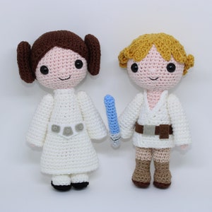 Puede incluir: Dos muñecos de ganchillo, uno con cabello castaño en moños laterales y un vestido blanco, el otro con cabello rubio y una túnica blanca, sosteniendo un sable de luz azul. Los muñecos están hechos a mano y tienen ojos negros y caras sonrientes.