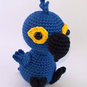 May include: A handmade crochet bird in shades of blue, yellow, and black. The bird has a round body, a black beak, and large black eyes. The wings and feet are also black. The bird is sitting upright, with its head tilted slightly.