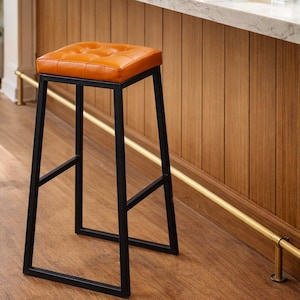 May include: A modern bar stool with a square, tufted orange seat and a black metal frame. The stool is set against a backdrop of a wooden wall and a marble countertop, suggesting a kitchen or bar setting.