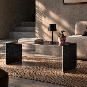 May include: A dark gray metal coffee table with a smooth, rectangular top and curved side supports. On the table sits a small, black, cylindrical battery-operated lamp, a stack of two light-colored books, and a small potted green succulent plant.