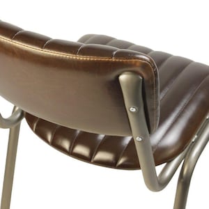 May include: Close-up of a brown leather chair with a curved back and seat. The chair has a metal frame with visible rivets. The leather is stitched with a quilted pattern, and the metal has a brushed finish.