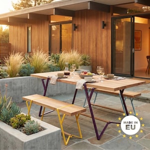 Può includere: Set da pranzo all'aperto con tavolo e panche in legno chiaro. Il tavolo ha gambe viola e le panche gambe gialle. Il tavolo è apparecchiato con piatti, bicchieri e una ciotola di frutta.