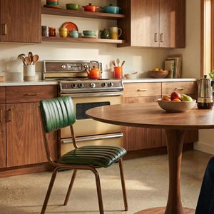May include: A retro kitchen with wooden cabinets, shelves, and a round table. A green leather chair is in front of the table. The kitchen includes a stove, a bowl of fruit, and various kitchenware.