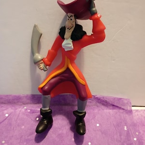 May include: A plastic figurine of a pirate character with a long dark mustache, wearing a red coat with yellow trim, purple pants, black boots, and a red hat with a grey feather. The figure holds a grey curved sword in one hand and touches the hat.