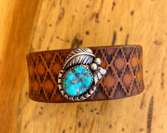 May include: A wide brown leather cuff bracelet featuring a repeating diamond pattern stamped into the surface. Centered on the leather is a silver ornament shaped like a feather and a small cluster of spheres holding an oval turquoise stone.