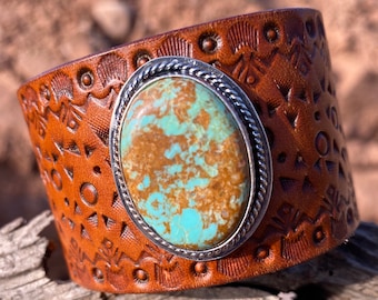 May include: A brown leather cuff bracelet with a silver setting and a turquoise stone. The leather is embossed with a geometric pattern.