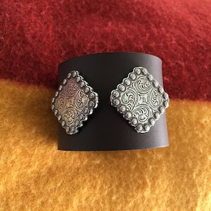 May include: A black leather cuff bracelet with two silver, diamond-shaped, engraved conchos.