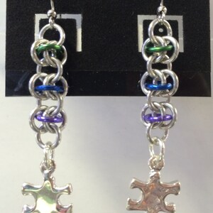 May include: A pair of silver earrings with a chain of silver rings with green, blue, and purple accents. Each earring has a silver puzzle piece charm hanging from the bottom.