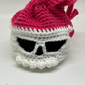 Pode incluir: Uma bolsa de caveira de crochê feita à mão em branco, preto e rosa choque. A bolsa apresenta um design de caveira com órbitas pretas e um fecho de cordão. A parte superior da bolsa é rosa choque e a caveira é branca.