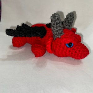 May include: A small, red crocheted dragon with black wings and gray horns. The dragon has a blue eye and is lying on its belly.