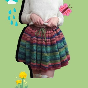 May include: A colorful striped crochet skirt with a brown drawstring waist. The skirt is worn by a person with a white fuzzy sweater and a yellow undershirt. The background is a green color with a rainbow, a cloud with raindrops, a butterfly, and a sun.