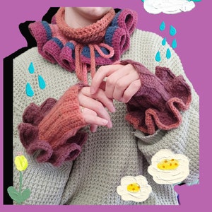 Puede incluir: Un suéter de punto gris con un cuello de ganchillo rosa y azul y calentadores de muñeca a juego. El cuello tiene un borde con volantes y los calentadores de muñeca tienen un borde con volantes.