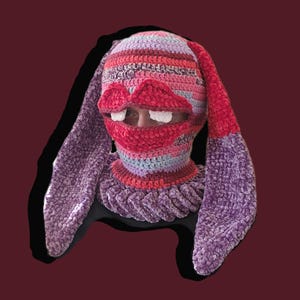 Puede incluir: Un pasamontañas de ganchillo hecho a mano con un diseño caprichoso. El pasamontañas presenta un patrón de rayas en tonos de rosa, rojo, morado y azul. Incluye labios rojos grandes con dientes blancos y largas orejas de conejo.