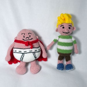 Peut inclure: Deux jouets en peluche crochetés. L'un est un personnage rond rose portant une cape rouge et des sous-vêtements blancs. L'autre est un personnage blond portant une chemise rayée verte et blanche et un short marron.