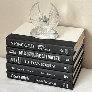 May include: A stack of books with black covers and white text, topped with a clear glass eagle figurine. The book titles include "STONE COLD", "MISUNDERESTIMATED", "13 BANKERS", "THE LAST DAYS", and "Don't Blink".