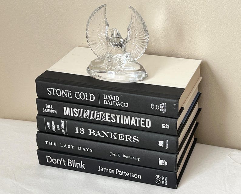 May include: A stack of books with black covers and white text, topped with a clear glass eagle figurine. The book titles include "STONE COLD", "MISUNDERESTIMATED", "13 BANKERS", "THE LAST DAYS", and "Don't Blink".