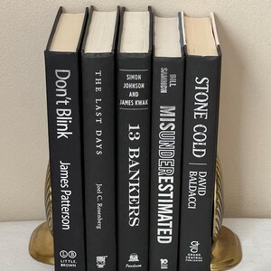 May include: Five hardcover books with black covers and white text are displayed. The titles include "Don't Blink," "The Last Days," "13 Bankers," "Misunderestimated," and "Stone Cold." The books are held by gold-colored bookends.