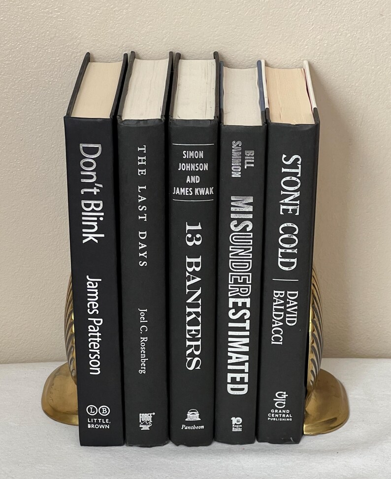 May include: Five hardcover books with black covers and white text are displayed. The titles include "Don't Blink," "The Last Days," "13 Bankers," "Misunderestimated," and "Stone Cold." The books are held by gold-colored bookends.