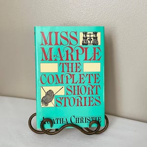 May include: A teal book titled "Miss Marple: The Complete Short Stories" by Agatha Christie. The book is displayed on a decorative, wrought-iron stand. The title is in red, with illustrations.
