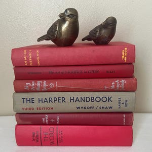 May include: Two bronze bird figurines rest atop a stack of vintage books. The books are primarily red, with titles like "An Actor Prepares" and "The Harper Handbook." The overall aesthetic is classic and decorative, suitable for home decor.