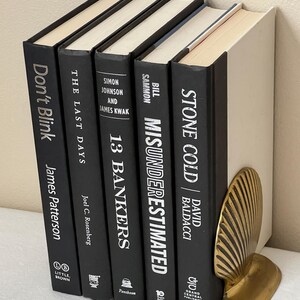 May include: A collection of hardcover books with black covers and white lettering, including titles like "Don't Blink" and "Stone Cold." A gold-colored seashell bookend supports the books. The books are arranged vertically, suggesting a bookshelf or display.