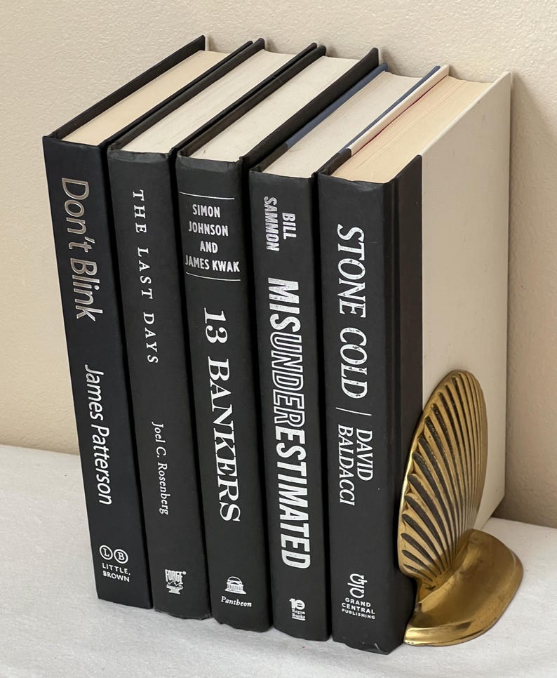 May include: A collection of hardcover books with black covers and white lettering, including titles like "Don't Blink" and "Stone Cold." A gold-colored seashell bookend supports the books. The books are arranged vertically, suggesting a bookshelf or display.