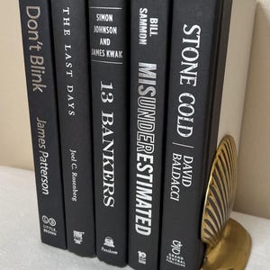 May include: A collection of black hardcover books with white text, including titles like "Don't Blink" and "Stone Cold." A gold-colored, leaf-shaped bookend supports the books. The books are arranged vertically.
