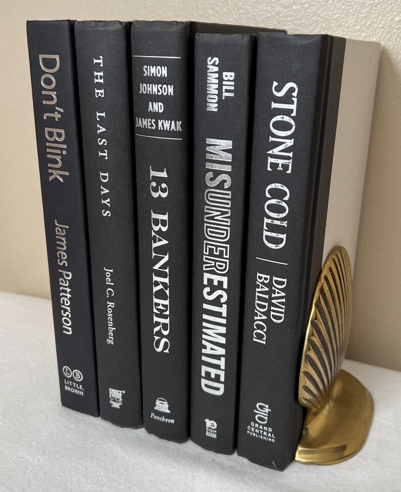 May include: A collection of black hardcover books with white text, including titles like "Don't Blink" and "Stone Cold." A gold-colored, leaf-shaped bookend supports the books. The books are arranged vertically.