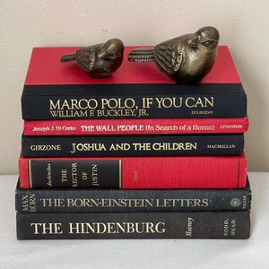 May include: A stack of vintage books with red and black covers, topped with two bronze bird figurines. The titles include "MARCO POLO, IF YOU CAN" and "THE HINDENBURG". The books are stacked on a white surface.
