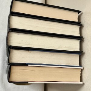 May include: A stack of five hardcover books with black spines and off-white pages. The books are arranged vertically, with the top book's pages facing up. The edges of the pages are a light tan color.