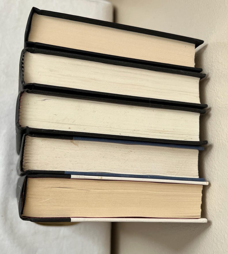May include: A stack of five hardcover books with black spines and off-white pages. The books are arranged vertically, with the top book's pages facing up. The edges of the pages are a light tan color.