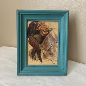May include: A framed artwork featuring a person with their head in their hands. The painting has a vintage aesthetic with warm tones and a distressed teal frame. The artwork is a portrait with a focus on the subject's face.