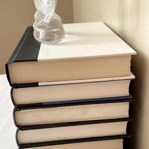 May include: A stack of five hardcover books with black spines and cream-colored pages. A clear glass sculpture of a horse is perched on top of the books. The books are stacked on a white surface.