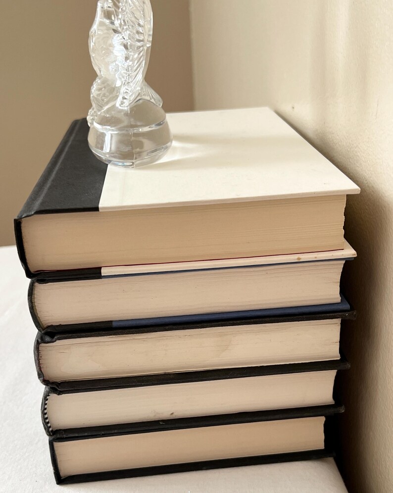 May include: A stack of five hardcover books with black spines and cream-colored pages. A clear glass sculpture of a horse is perched on top of the books. The books are stacked on a white surface.