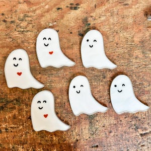 May include: Six white ghost-shaped ornaments with black eyes and mouths, some with red hearts. The ghosts are on a wooden surface.