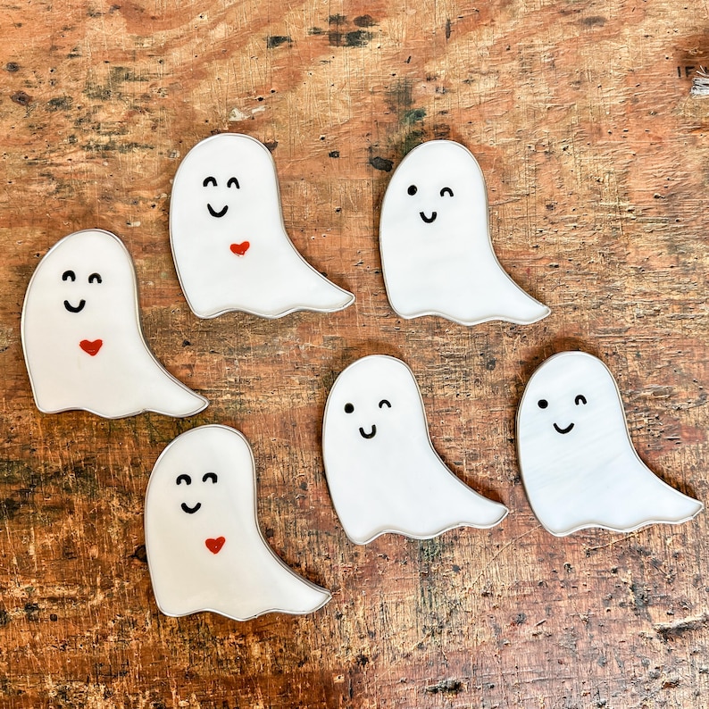 May include: Six white ghost-shaped ornaments with black eyes and mouths, some with red hearts. The ghosts are on a wooden surface.