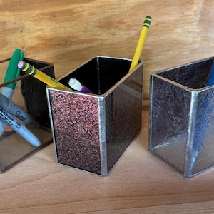 May include: Three decorative desk organizers made of glass and metal. The organizers are rectangular and hold pens, pencils, and markers. One has a textured maroon finish, another has a silver finish, and the third has a clear finish.
