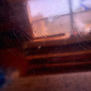 May include: A close-up shot of a reflective, copper-colored surface. The surface has scratches and imperfections, with reflections of a window and other objects visible. The overall effect is a blend of warm and cool tones.