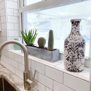 May include: A white ceramic vase with a speckled brown glaze sits on a windowsill with a planter box filled with succulents and cacti. The windowsill is tiled with white subway tiles.