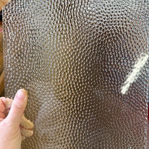 May include: A sheet of textured glass with a repeating pattern of small, rounded bumps. The glass is held by a hand, and the light reflects off the surface, creating highlights and shadows. The glass appears to be clear or slightly tinted.