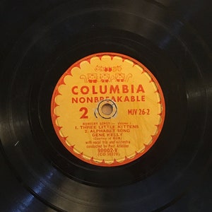 Op de afbeelding: Een zwarte vinylplaat met een geel en oranje label. Het label zegt "Columbia Nonbreakable 2 MJV 26-2" en bevat de songtitels "Three Little Kittens" en "Alphabet Song" van Gene Kelly.
