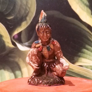May include: A small, vintage figurine of a Native American man in a squatting position. The figurine is painted in shades of brown, with colourful accents on the headdress and clothing. The background is a blurred image of green and yellow leaves.