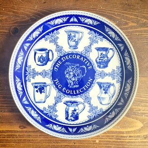 May include: A round, decorative plate with a blue and white design. The plate features a central medallion with the text "The Decorative Jug Collection" and illustrations of various jugs. The border has a repeating pattern.