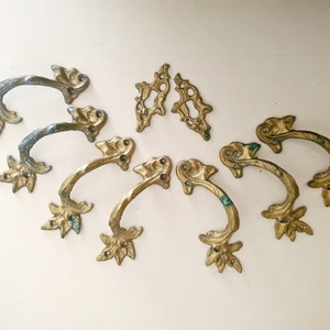 Może przedstawiać: Osiem ozdobnych, złoconych metalowych uchwytów do szuflad z detalami w kształcie liści. Uchwyty mają zakrzywiony kształt i są przeznaczone do użytku w meblach.
