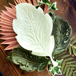 Puede incluir: Tres fuentes de cerámica con forma de hoja en blanco y verde oscuro. La fuente blanca está en la parte superior, con un diseño detallado de venas de hoja. Las fuentes están dispuestas sobre una superficie de madera con hojas de palma secas y vegetación.