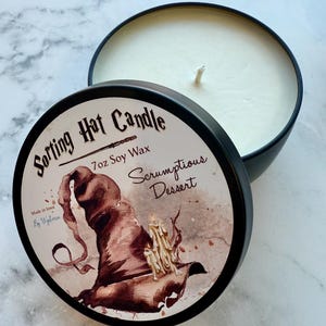 May include: A black tin candle with a white wax interior and a wick. The lid features a brown Sorting Hat illustration with the text "Sorting Hat Candle" and "Scrumptious Dessert". The candle is 7oz and made with soy wax.
