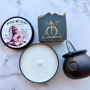 May include: An assortment of themed items, including a candle in a black tin with the text "Sorting Hat Candle", a dark gray soap with a gold symbol, and a small black cauldron. The candle has a white wax.