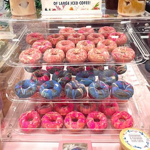 May include: A display case filled with colorful donuts. The top shelf features pink and red frosted donuts. The middle shelf has blue and black frosted donuts. The bottom shelf has pink frosted donuts. The donuts are arranged on clear plastic shelves.